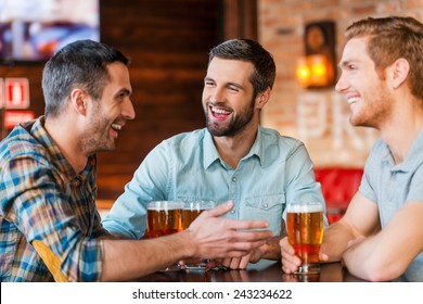 Meeting With The Best Friends. Three Happy Young Men In Casual Wear Talking And Drinking Beer While Sitting In Bar Together