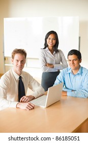 Meeting Of Diverse Team Of Business People In Bright Conference Room, Asian Female, Latino Man And Caucasian Male Sitting Around Table With Laptop Smiling, Looking At Camera. Vertical Copy Space