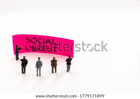 Meeting with miniature figurines posed as business people standing around post-it note with Social Unrest handwritten message in background, minimalist abstract concept with focus on text
