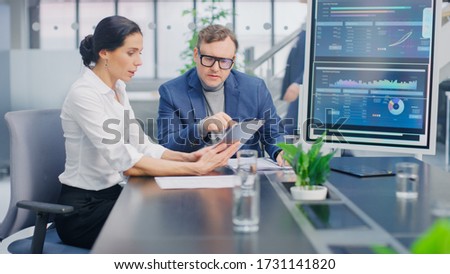 In the Meeting Room Female Executive Shows Digital Tablet Computer to Creative Venture Capitalist, They Discuss Statistics and Investment Capital. Busy Corporate Office with Businesspeople Working