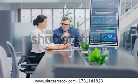 In the Meeting Room Female Executive Shows Digital Tablet Computer to Creative Venture Capitalist, They Discuss Statistics and Investment Capital. Busy Corporate Office with Businesspeople Working