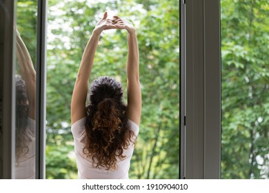 Meeting summer day. Back rear view of happy young woman stand in balcony door stretch hands enjoy weekend have pleasure. Calm beautiful female do morning exercises practice yoga by opened large window - Powered by Shutterstock