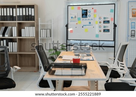 Meeting table with stacked papers, documents, and a red mug, in a modern office setting with a whiteboard displaying charts, graphs, and sticky notes, suggesting a collaborative workspace