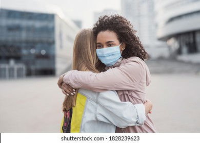 Meeting of two happy friends hugging in the street wearing protective face mask. Multiethnic young women embrace each others in a city during corona virus pandemy - Powered by Shutterstock