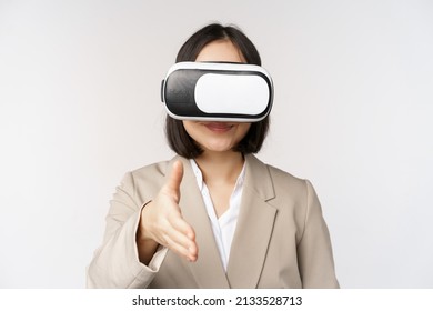 Meeting in vr chat. Asian businesswoman in virtual reality glasses, extending hand for handhshake with business partner, greeting someone, standing over white background - Powered by Shutterstock