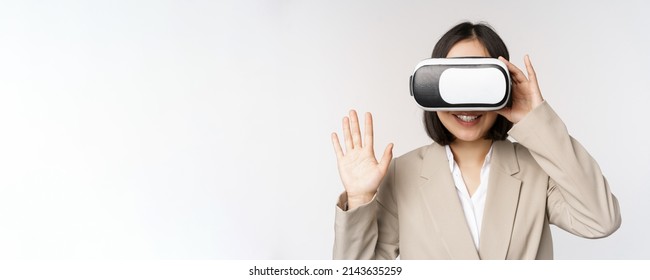 Meeting in vr chat. Asian businesswoman in virtual reality glasses, raising hand and saying hello, greeting someone, standing over white background