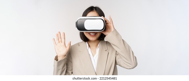 Meeting in vr chat. Asian businesswoman in virtual reality glasses, raising hand and saying hello, greeting someone, standing over white background