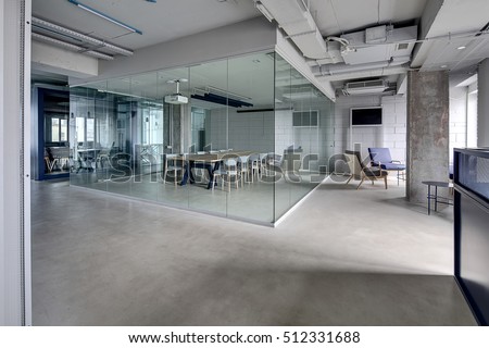 Meeting zone in the office in a loft style with white brick walls and concrete columns. Zone has a large wooden table with gray chairs and glass partitions. Above the table there is a projector. 