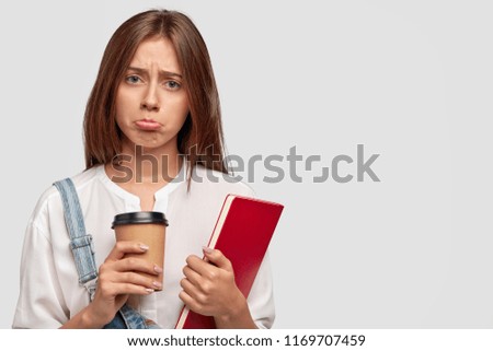 Melancholic green eyed displeased beautiful schoolgirl purses lips from displeasure, holds disposable cup of drink and red book, doesnt want to study, dressed in white shirt and denim dungarees