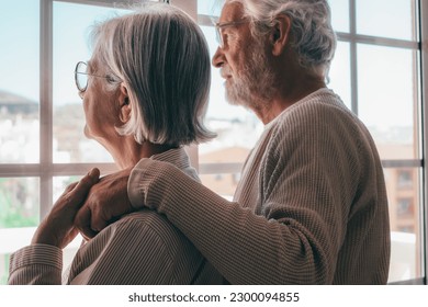 Melancholic senior family couple of Caucasian people hugging each other with love, elderly man and woman with glasses support each other in a moment of sadness looking out the window - Powered by Shutterstock