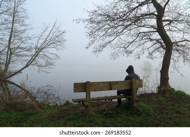 Melancholy Emotions Concept: Depressed Man Grieving On A Bench At A Lake On A Foggy Day.