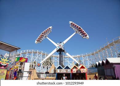 Melbourne, Australia: March 18, 2017: A Ride Is In Full Swing At Melbourne's Luna Park. The Historic Amusement Park Is Located On The Foreshore Of Port Phillip Bay In St Kilda. People Walk By. 