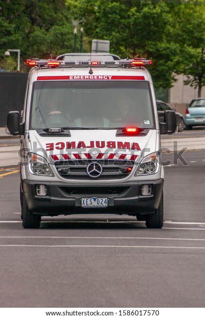 Melbourne,\
Australia - November 16, 2009: Frontal view on white and red\
ambulance emergency Mercedes van riding with flashing lights in\
street. Green foliage and cars in\
back.