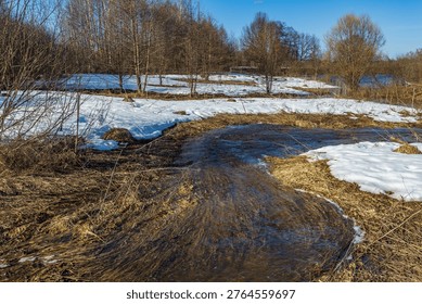Melted snow flows through a bare, sunlit field. Dried grass and branches frame the shallow stream