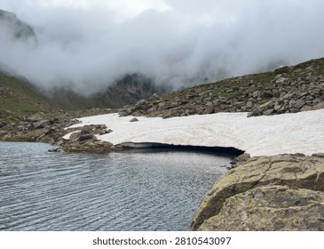 Melting snow forms a dramatic overhang above a glacial lake fed by snowmelt at Tristaina Lakes, Andorra. Misty clouds shroud rocky mountain slopes of the Pyrenees