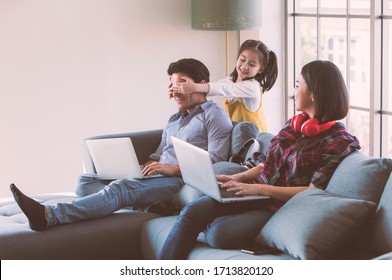 Members Of Diverse Family, Caucasian Father And Asian Mother Using Laptop Computer Notebook To Work From Home, Young Cute Little Girl Daughter Come To Kidding Her Dad By Closing His Eyes With Hands.