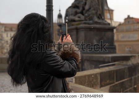 Memory keeper photographing bridge statue details, hands holding smartphone close to bronze sculpture, river and historic buildings softened in background, textured masonry