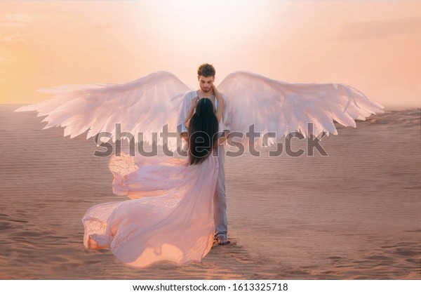 Men angel hugs young woman. Long dark hair dress\
silk fabric flying flutter wind. costume white wings. Bright color\
peach pink yellow sunset desert sky. Shoot back rear view turned\
away without face