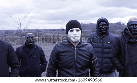 Men in black jackets and masks. Footage. Criminal gang in black plastic masks and leader in white mask on background of cloudy sky. Masked social criminal gang