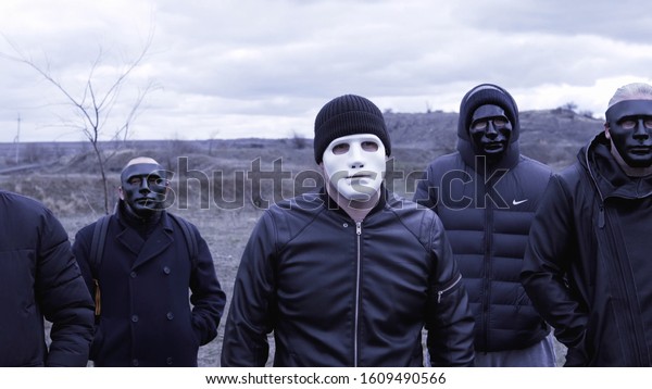 Men in black jackets and masks.\
Footage. Criminal gang in black plastic masks and leader in white\
mask on background of cloudy sky. Masked social criminal\
gang