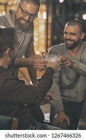 Men Clinking Crystal Glasses With Alcohol, Looking At Each Other And Smiling. Friends Having Fun, Relaxing In Bar. Men Celebrating And Drinking Delicious Whiskey Or Scotch.