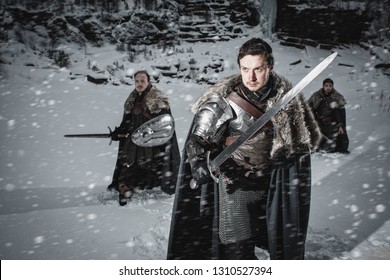 Men Dressed In Medieval Armor And Raincoats With Swords In Winter In The Mountains Under Heavy Snow Blizzard. Fantasy Group Of Warrior Knights In Winter Snowy Landscape