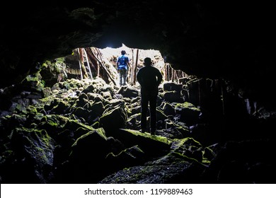 Men exploring a cave made by lava eruptions in a rainforest, between rocks and moss.