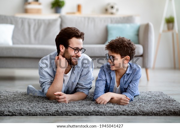 Men family.\
Cheerful father and son lying on floor carpet and lookig at each\
other, wearing eyeglasses and smiling, spending time together at\
home, enjoying weekend. Fatherhood\
concept