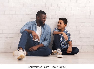 Men Family. Portrait Of Happy African American Father And Son Sitting Together On Floor Against White Brick Wall And Lookig At Ech Other, Copy Space
