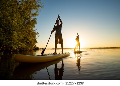 Men, friends sail on a SUP boards in a rays of rising sun. Stand up paddle boarding - awesome active recreation in nature. Backlight, wide angle. 