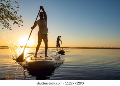 Men, friends sail on a SUP boards in a rays of rising sun. Stand up paddle boarding - awesome active recreation in nature. Backlight. 