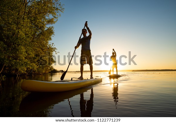Men, friends sail on a SUP boards in a rays\
of rising sun. Stand up paddle boarding - awesome active recreation\
in nature. Backlight, wide angle.\
