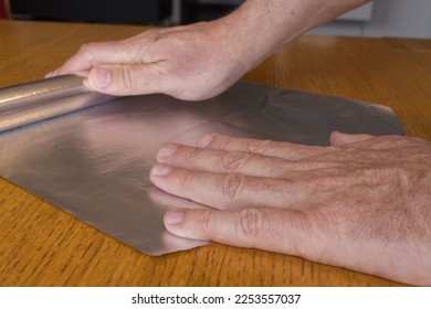 Men hands roll off the aluminum foil for household use on a wooden surface.   - Shutterstock ID 2253557037