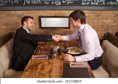 men laugh together while sitting in cafe. two man holding hands and greeting each other at table