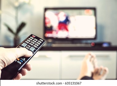 Men with the remote control front of the television. Hand with remote control directed on the TV. A man is relaxing and watching sports on TV.