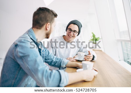 Men are sitting in front of the table and near window. Happy young guys are looking at each other and talking about girls.
