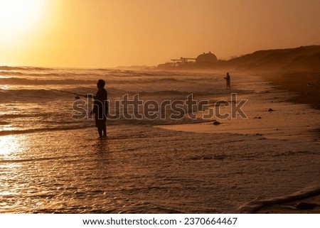 Men surf fishing among ocean waves at sunset with a power plant in the background 