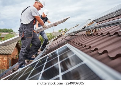 Men technicians carrying photovoltaic solar moduls on roof of house. Builders in helmets installing solar panel system outdoors. Concept of alternative and renewable energy.