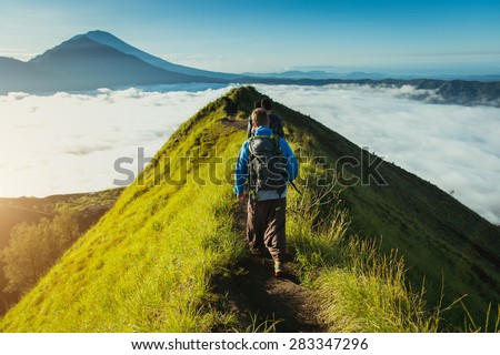 Men walk along the hill with backpacks and with white clouds and peak of volcano on background