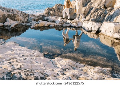 Men walk holding hands along a rocky seashore reflected in a puddle - Powered by Shutterstock