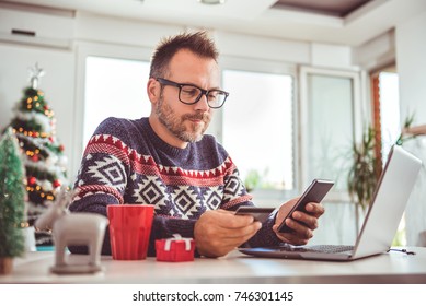 Men wearing blue sweater and eyeglasses holding credit card and using laptop at home office during christmas holidays