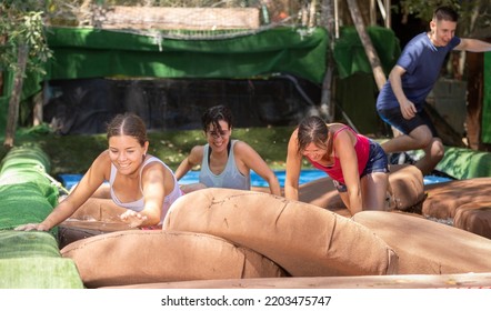 Men And Women Jumping On Floating Platforms To Reach Finish Of Water Obstacle Course. Family Resting In Amusement Park In Summertime.