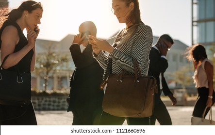 Men and women using mobile phone while commuting to office on a crowded street with sun flare in the background.
