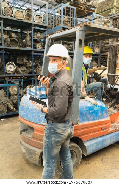 Men work\
together, wear safety facemask, use walkie-talkie. Caucasian\
engineer man wear safety facemask and use walkie-talkie while Asian\
man driving forklift in\
factory-warehouse