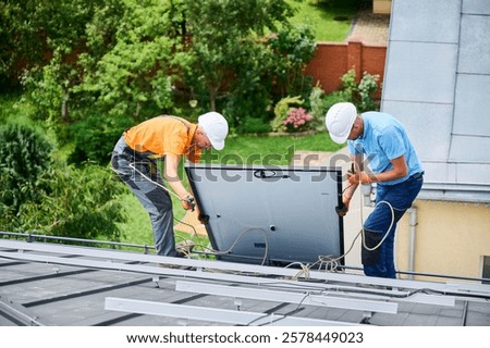Men workers installing solar panel system on roof of house. Electricians in helmets lifting up photovoltaic solar module with help of ropes outdoors. Concept of alternative and renewable energy.