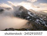 Menacing clouds and fog rise from the valley to the ridge as the sun sets behind the peaks of Dachstein from mount Sinabell, Schladming, Austria