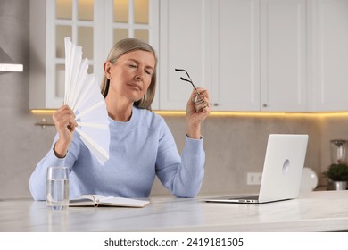 Menopause. Woman waving hand fan to cool herself during hot flash at table in kitchen - Powered by Shutterstock