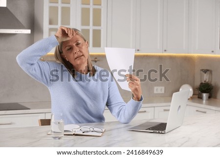 Menopause. Woman waving paper sheet to cool herself during hot flash at table in kitchen