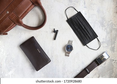 Men's Accessories During Quarantine. Brown And Blue. Flatlay On Gray Concrete. Black Medical Mask, Watch, Wallet, Briefcase And Belt.