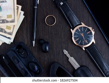 Men's Accessories On A Dark Wooden Background . The Concept Of Success And Business. Wrist Watch, Pen, Perfume, Headphones, Ring, Car Keys, Money And Phone. Flatlay. 